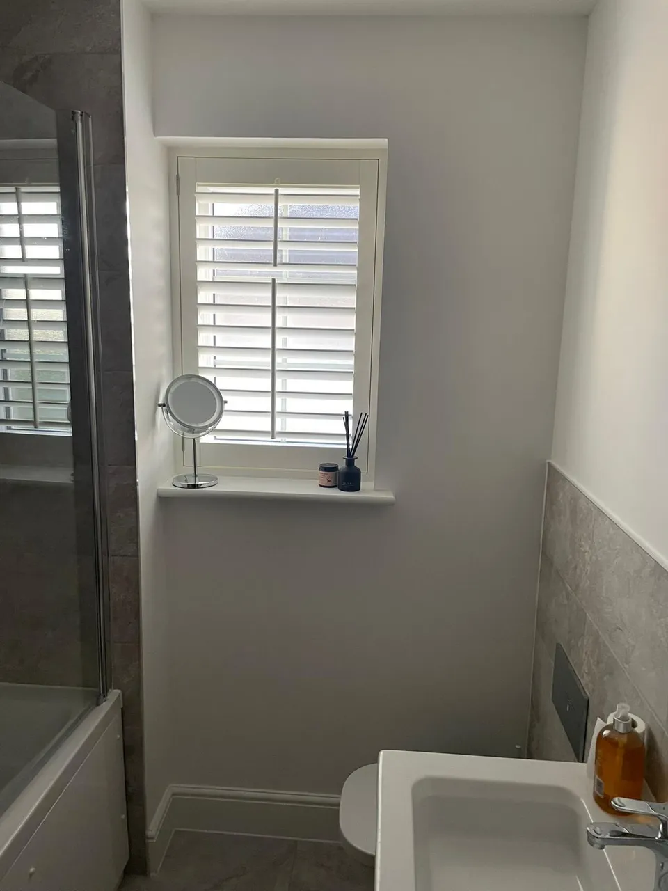 White plantation shutters on a small frosted window in a bathroom with a basin and bath