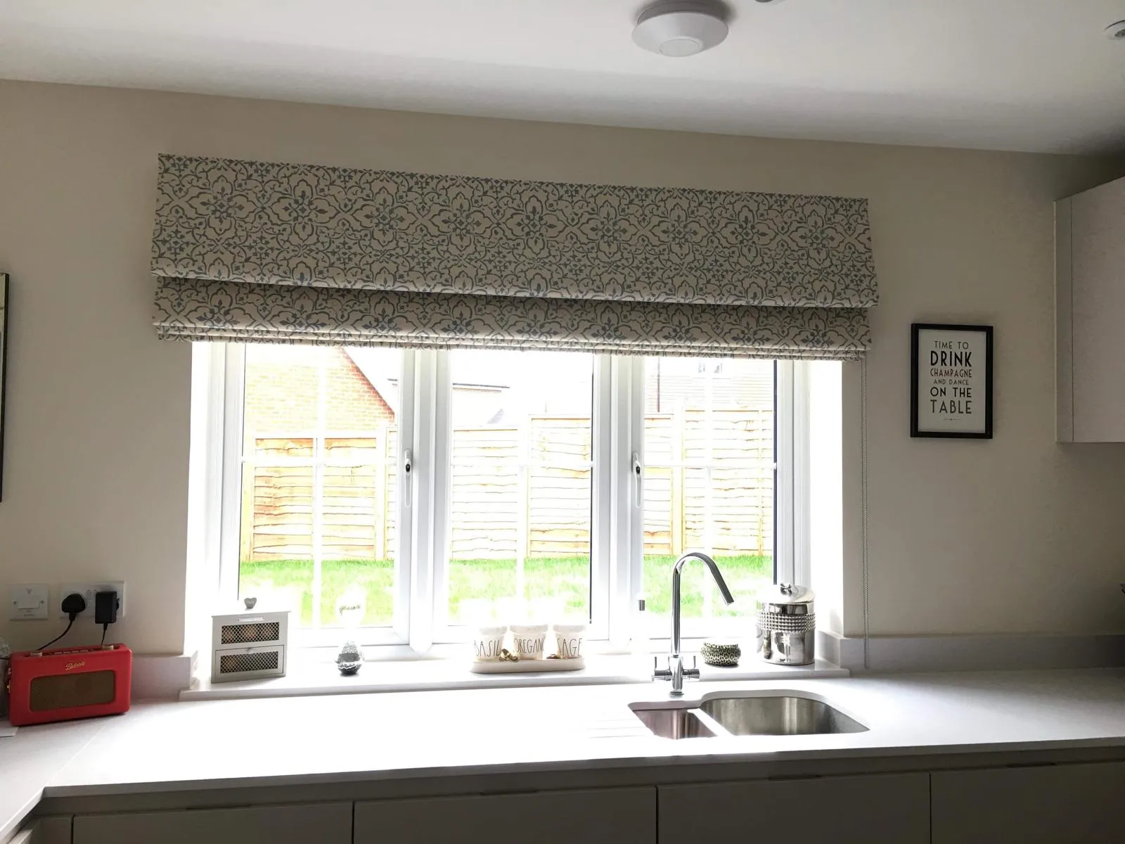 Patterned Roman blind above a kitchen sink window looking onto a fenced garden